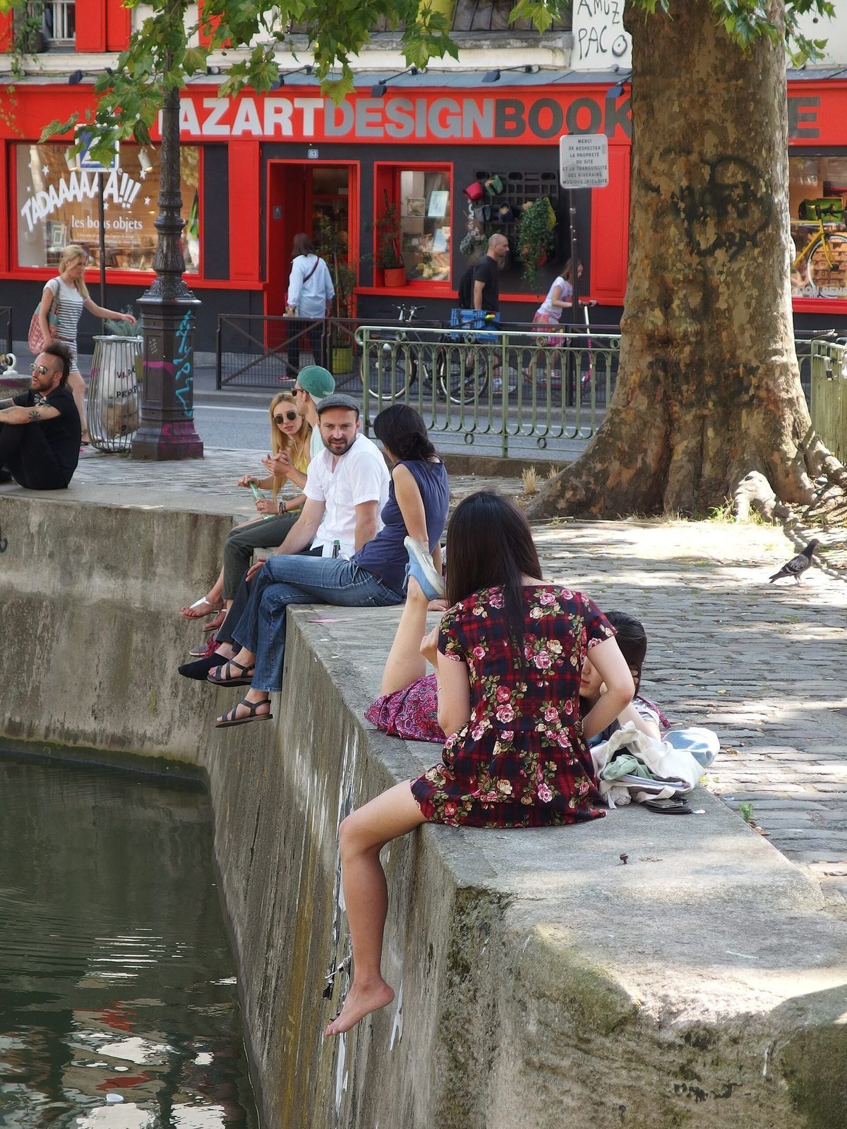 Folk som sitter tett langs kanten av Canal Saint-Martin en sommerdag, med trær og bygårder bak