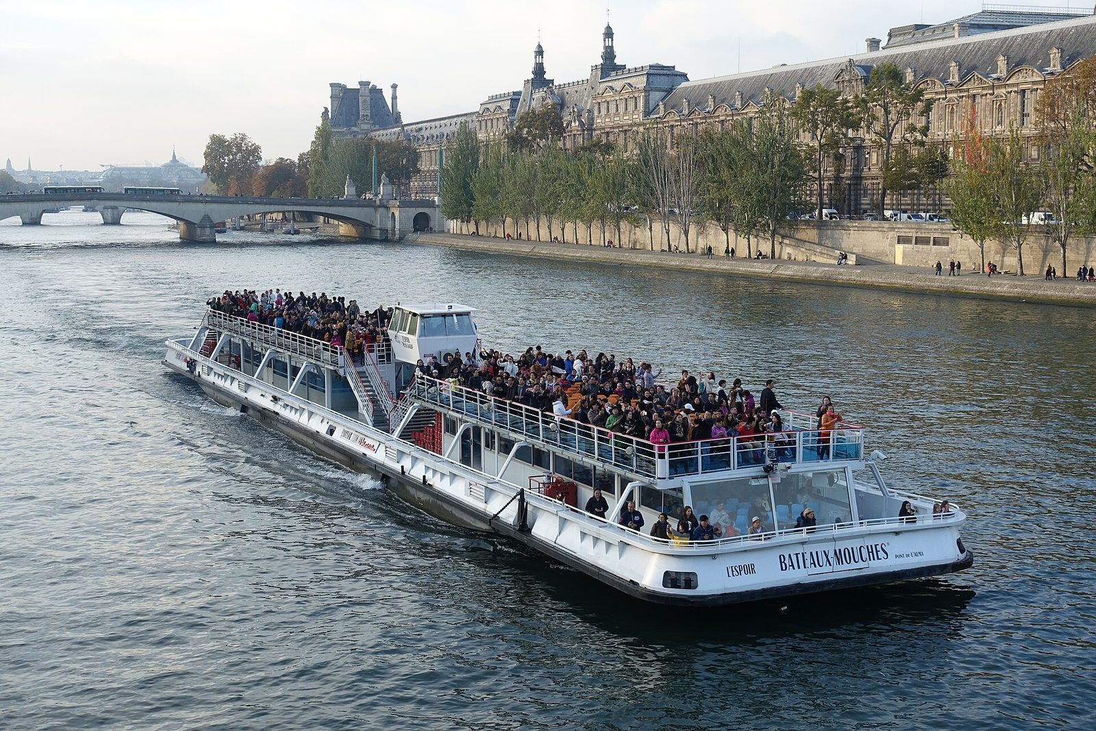 Fullastet turistbåt på Seinen med passasjerer på åpent dekk, og Louvre-fløyen langs kaien bak