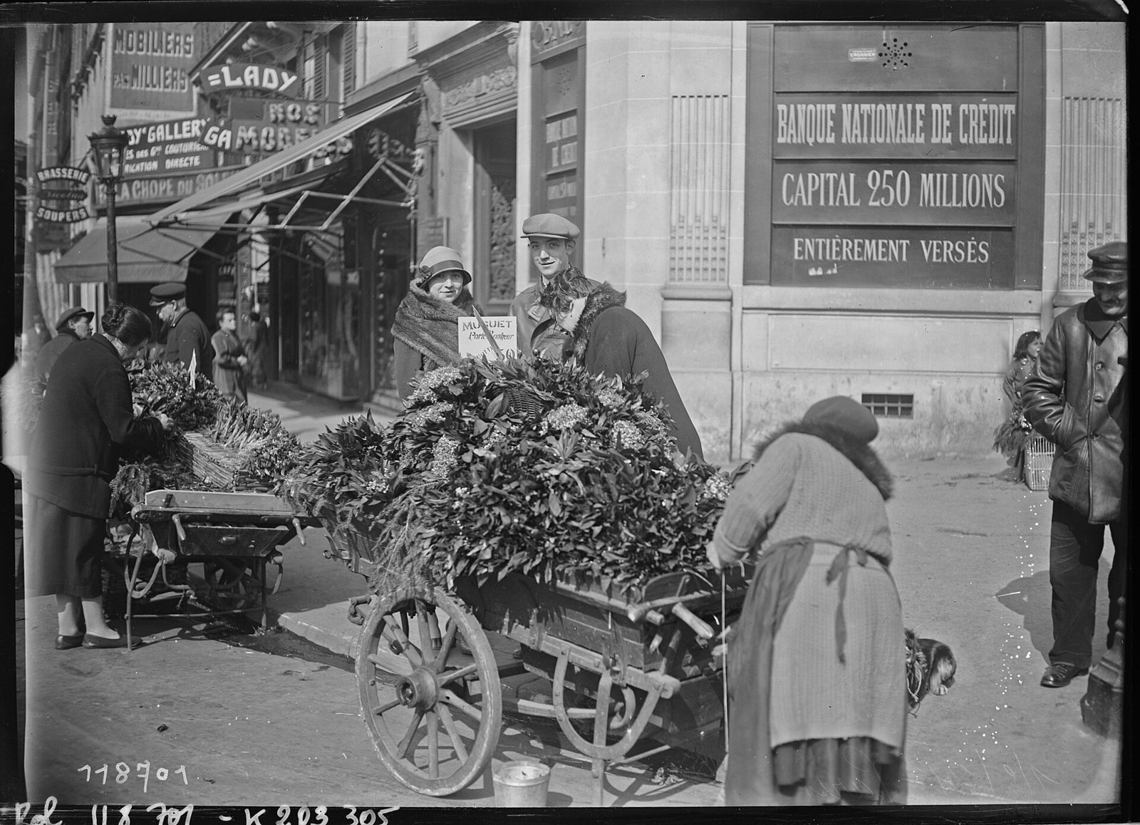 Selgere med håndkjerrer fulle av liljekonvaller på et gatehjørne i Paris, med skilt merket «Muguet»