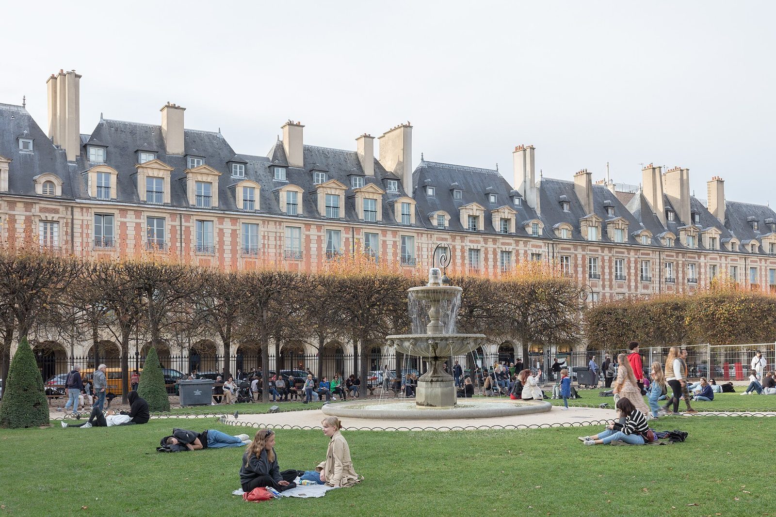 Folk sitter på plenen på Place des Vosges foran de røde teglpaviljongene en dag sent i oktober