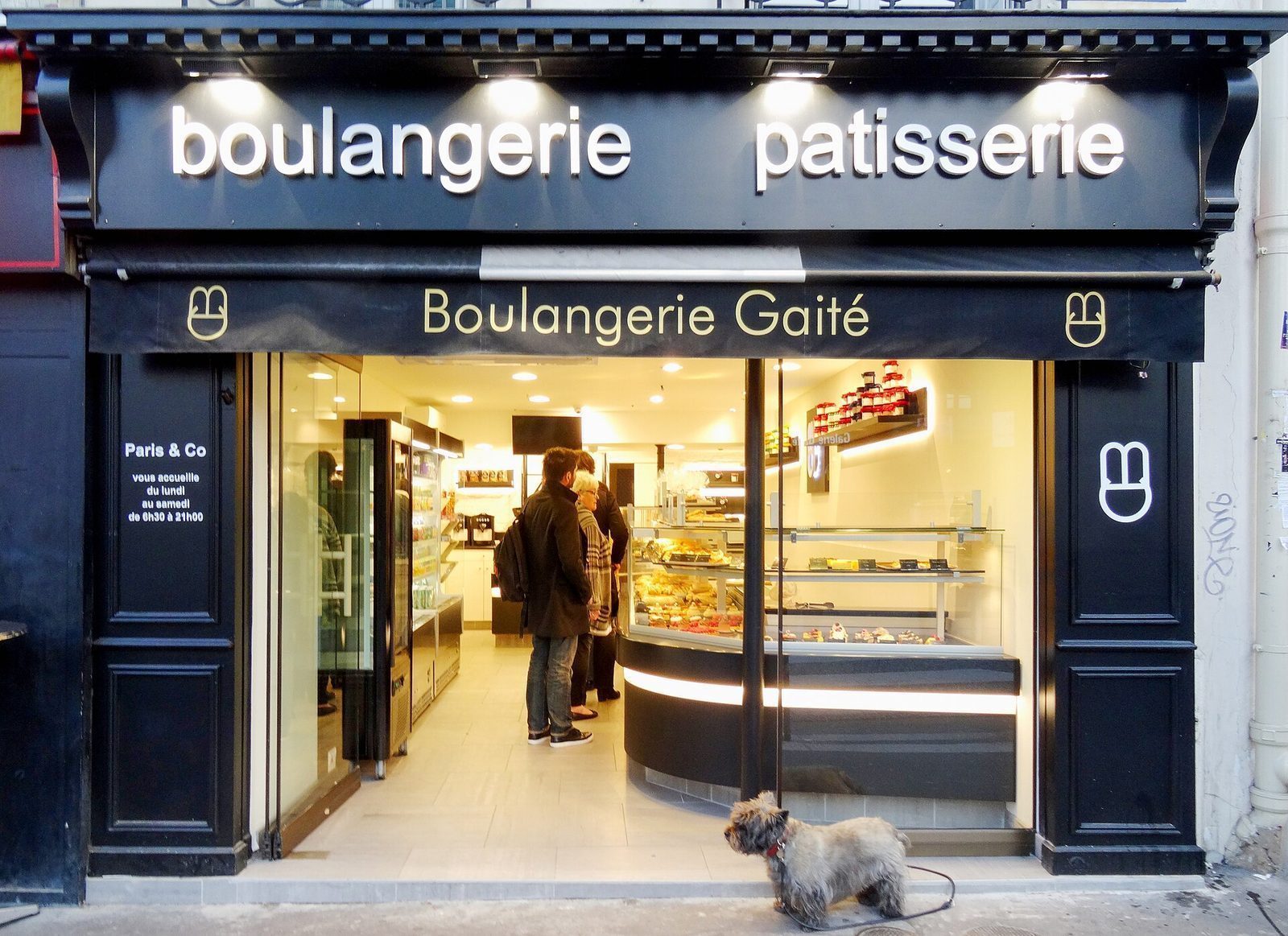Liten hund i bånd som venter utenfor en parisisk boulangerie mens eieren står i køen inne