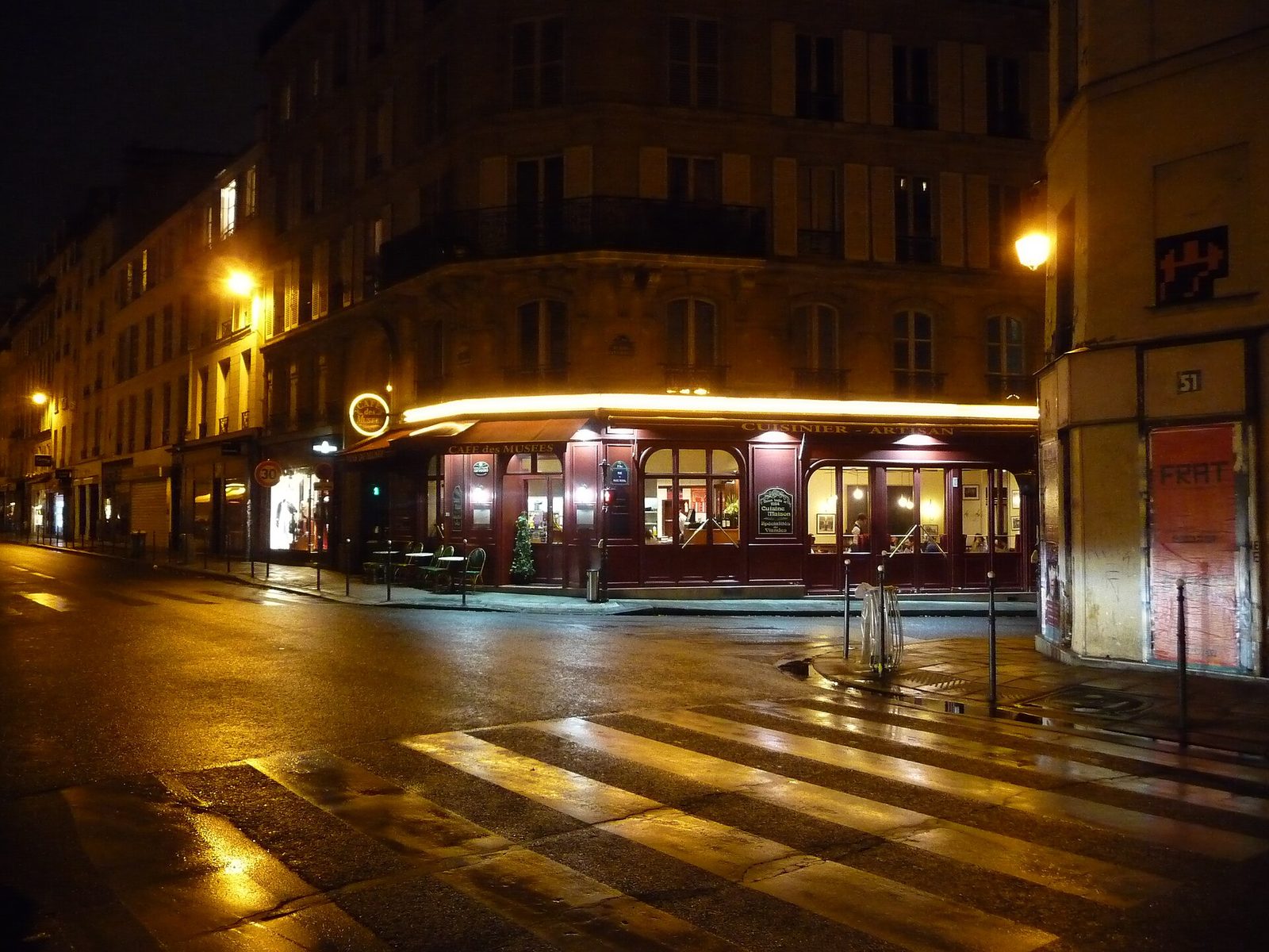 Opplyst hjørnekafé i Paris en kveld, med varmt lys ut i gaten og våt asfalt som speiler gatelyktene