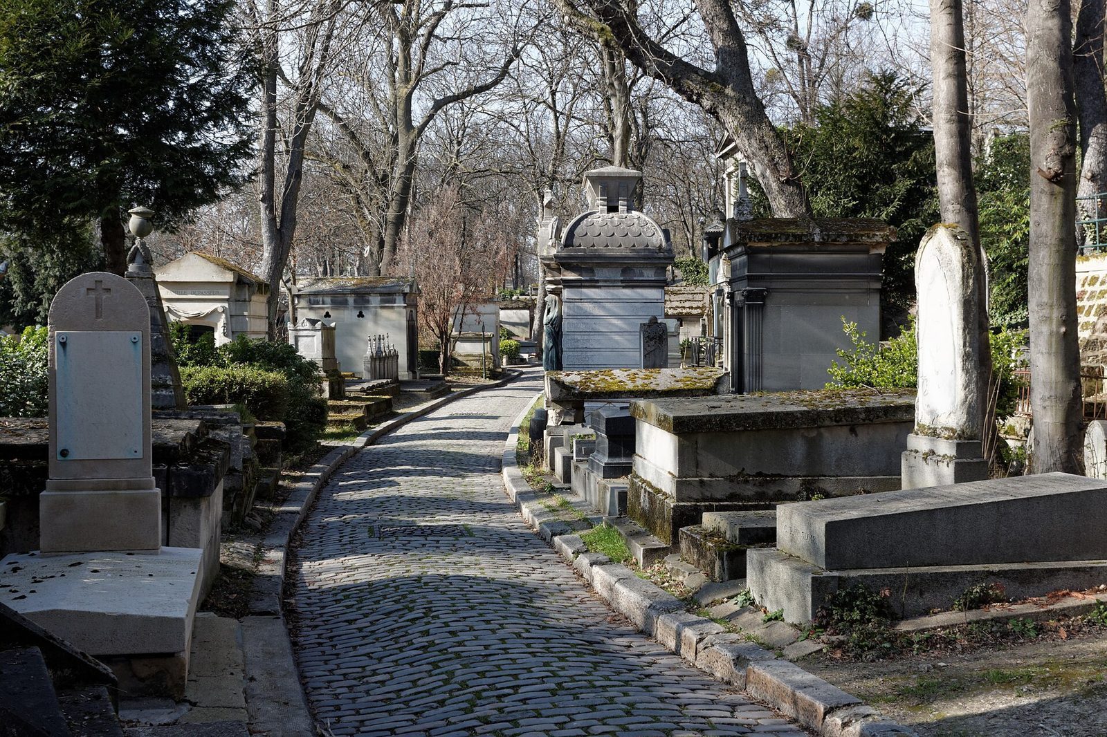 Brosteinsbelagt gravgate med mosegrodde gravkapeller og gamle trær på Père-Lachaise