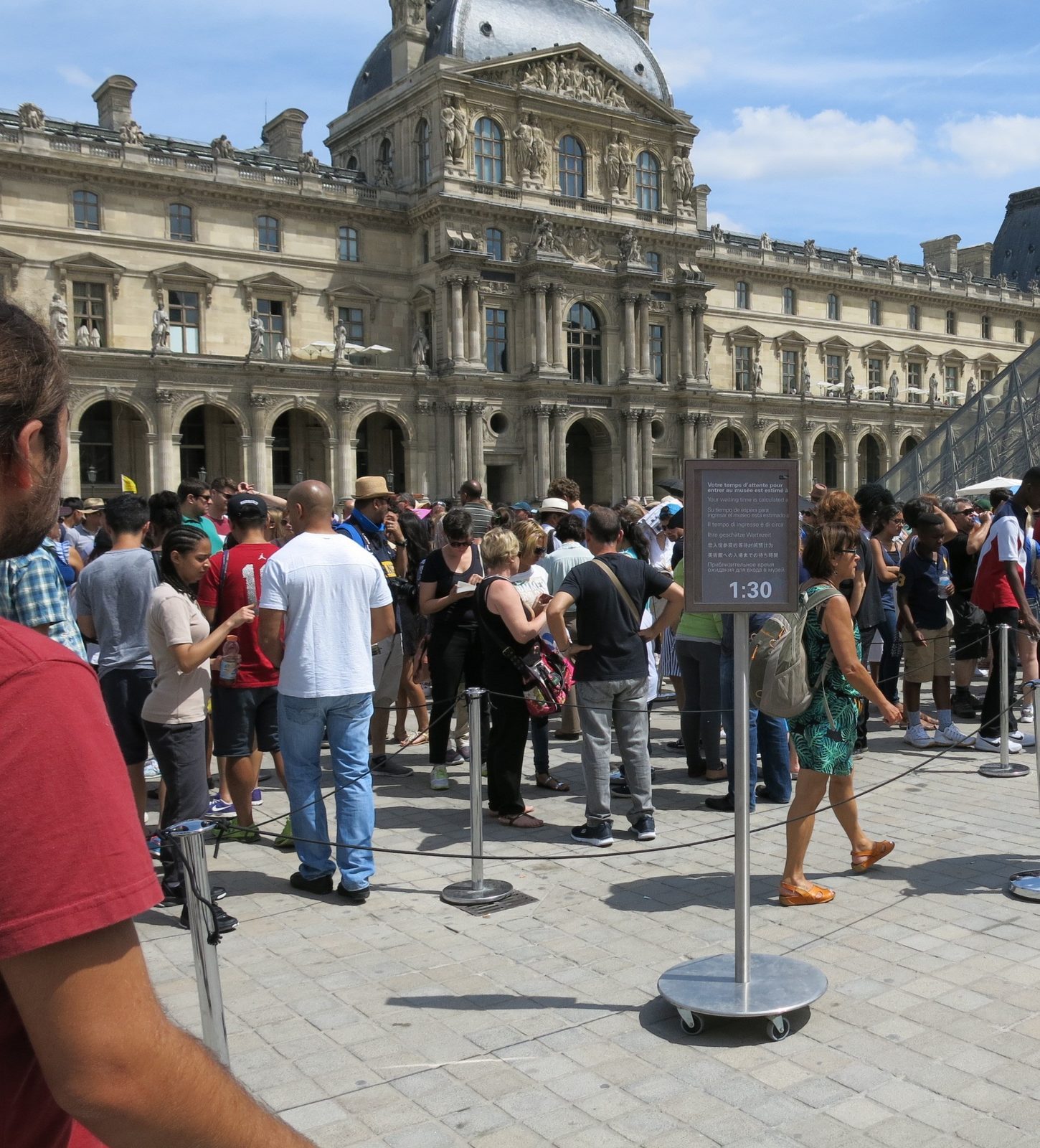 Tett kø av besøkende bak sperrebånd på plassen foran Louvre, med en skjerm som viser ventetid 1:30