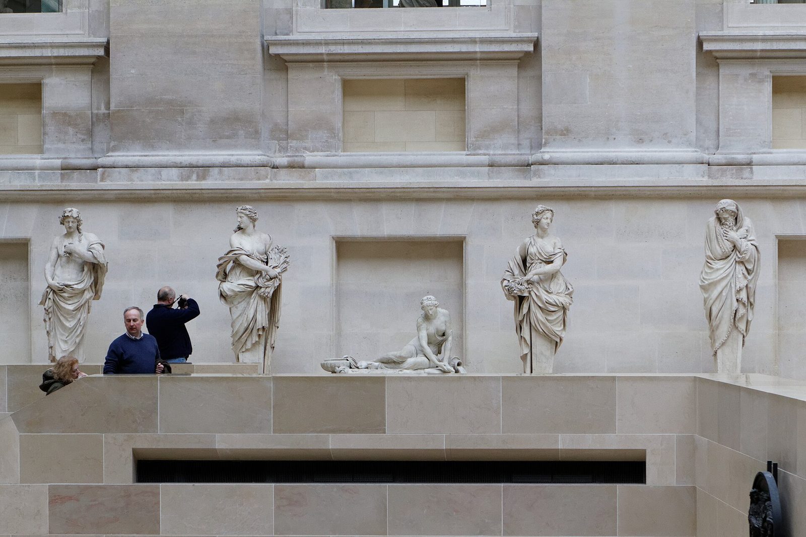 Skulpturer langs veggen i Cour Puget i Louvre, med bare tre besøkende ved rekkverket