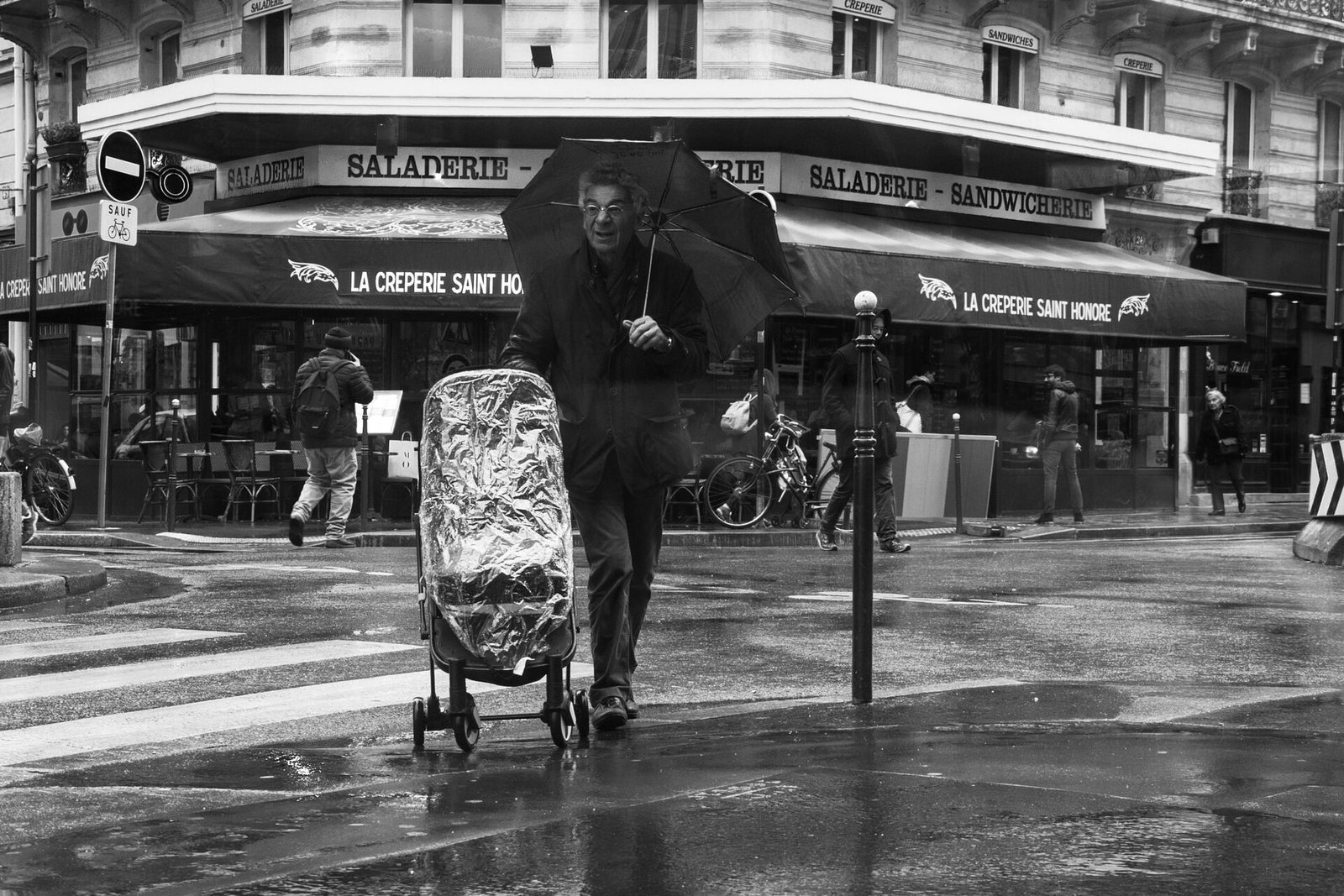 Svart-hvitt bilde av en fotgjenger med paraply som krysser en våt brosteinsgate i Paris